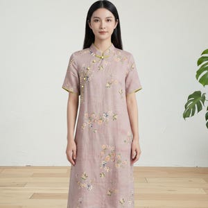 May include: A dusty rose-colored dress with a mandarin collar and short sleeves. The dress features a floral pattern and is trimmed with green piping. The dress has a button closure and is knee-length.