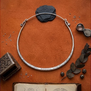 May include: A silver-toned metal torque necklace featuring a circular band with intricate engraved patterns along the lower curve and hook-style closures at the top, presented as a rigid collar piece with a polished finish and detailed decorative etchings.