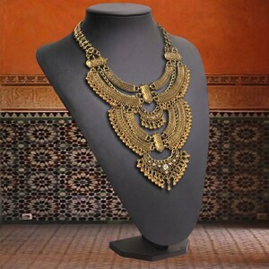 Make a bold impact with this antique gold bib necklace, featuring intricate engravings and resin stones designed for high-drama festival looks. Whether you are channeling a desert queen or elevating a bohemian outfit, this layered piece sits perfectly against the collarbone to command attention. Inspired by traditional nomadic adornments, this necklace uses weightier textures and a multi-tiered design to create movement and sound as you move.