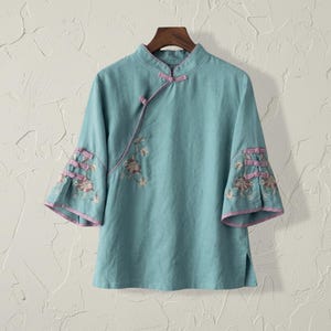 May include: A light blue, short-sleeved top with a mandarin collar and floral embroidery. The top features pink trim on the sleeves and neckline, and a side closure with fabric buttons. The garment is displayed on a wooden hanger.