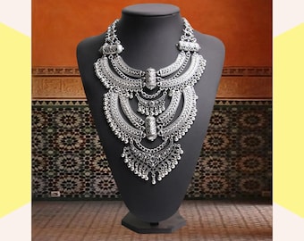 May include: A silver-toned statement necklace with an ornate, layered design. The necklace features multiple crescent-shaped elements and dangling pendants, displayed on a black mannequin. The background includes a patterned wall.