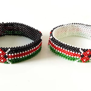 May include: Two beaded bracelets. One bracelet is black, red, white, and green. The other bracelet is white, black, red, and green. The bracelets are made of small beads and are likely handmade. The bracelets are on a white surface.