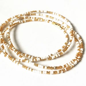 May include: A white and gold beaded waist chain. The chain is made of small, round beads that are strung together in a repeating pattern of white and gold.