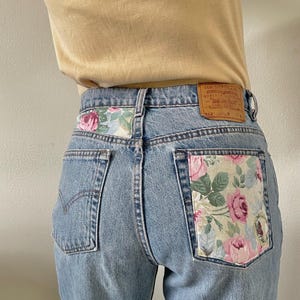 May include: Light blue denim jeans with a floral fabric patch on the back pocket. The patch features pink and green roses. The jeans have a Levi's label on the back.