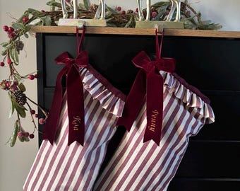 Op de afbeelding: Twee kerstsokken met een bordeauxrood en wit verticaal gestreept patroon en een gerimpelde bovenrand. Elke sok is versierd met een donkerrode fluwelen strik en een gepersonaliseerd naamplaatje met de namen Koa en Penny erop.