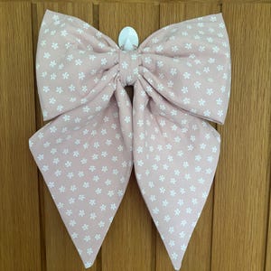 May include: A large fabric bow in a soft dusty pink colour featuring a repeating pattern of small white four-petalled flowers. The bow has two wide top loops and two long, pointed tails hanging downwards, secured by a central fabric knot.