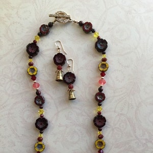 May include: A long necklace with dark red flower-shaped beads, yellow and clear beads, and a silver clasp. Matching earrings with dark red flower-shaped beads and small gold bells.