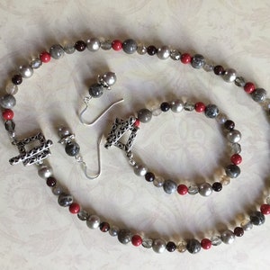May include: A set of jewelry featuring a necklace, bracelet, and earrings. The necklace and bracelet are made with grey, red, and brown beads. The earrings are silver with a filigree design.