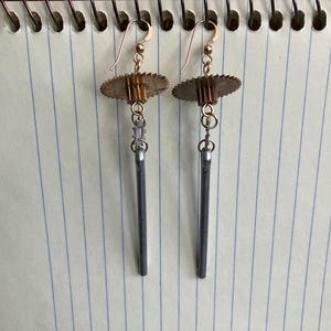 May include: A pair of steampunk-style earrings. Each earring features a gold-colored hook, a small gear-shaped element, a silver-colored chain, and a long, dark gray, rod-shaped pendant. The earrings are displayed on a lined paper background.