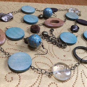 May include: A long necklace with a variety of wooden beads in shades of brown and blue. The necklace features a large, round, brown wooden bead with a hole in the center. The necklace also includes a pair of earrings with a small, round, blue wooden bead and a metal hook.