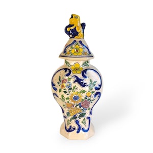 Könnte beinhalten: Dekorative Keramikvase mit Deckel, mit cremefarbenem Sockel und aufwendigen Blumendesigns in Gelb, Blau und Rot. Der Deckel ist mit einer gelben Vogelfigur gekrönt. Die Vase hat eine klassische, elegante Form.
