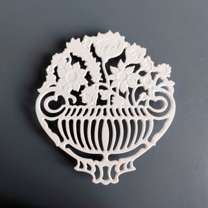 May include: White ceramic ornament shaped like a vase with a floral design. The vase has a scalloped rim and a decorative base. The flowers are stylised and include tulips, roses, and other blooms.