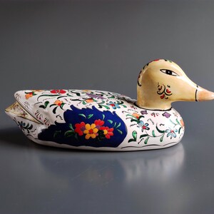 Puede incluir: Una figurita de pato de cerámica pintada a mano con una base blanca y diseños florales coloridos. El pato tiene un pico amarillo y un ala azul y blanca.