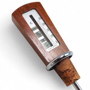 May include: A wooden wine thermometer with a silver metal ring and a cork base. The white display features a temperature scale in Celsius and labels for various beverages including Cognac, Rot Wein, Dessert Wein, Weiss Wein, Sekt, and Brannt Wein.