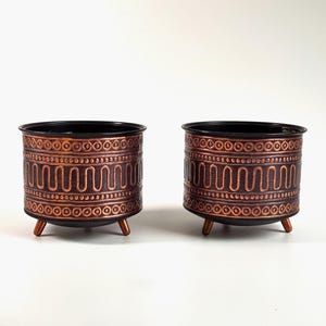 May include: Two copper-colored metal planters with black interiors and decorative patterns. Each planter has three short legs.