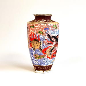 May include: A decorative vase with a hexagonal shape, featuring a detailed Japanese-style design. The vase has a maroon rim and base, with a colorful illustration of a dragon, waves, and other elements in red, blue, and gold.