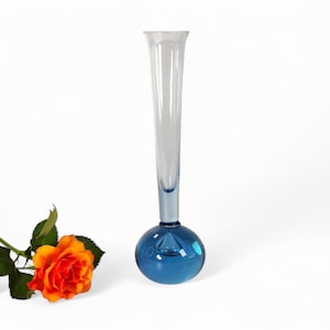 May include: A tall, clear glass vase with a blue, round base. The vase is narrow at the top and widens towards the bottom. A single orange rose is lying on the white surface next to the vase.