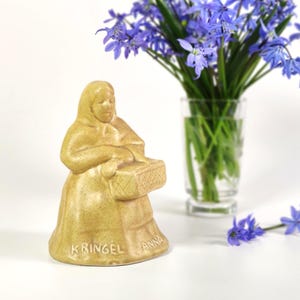 Può includere: Una statuetta in ceramica di una donna che tiene un cesto. La statuetta è dipinta in un colore marrone chiaro e ha le parole "KRINGEL ANNA" sulla base.