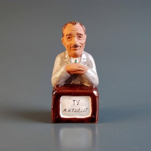 May include: A ceramic figurine of a man with a moustache, wearing a grey suit, sitting behind a brown television set. The TV has a white screen with the words "TV Aktuell" in black lettering.