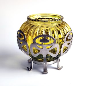 Puede incluir: Un portavelas de vidrio amarillo con una base de metal con un diseño decorativo. El portavelas tiene una textura acanalada y se apoya en tres patas.