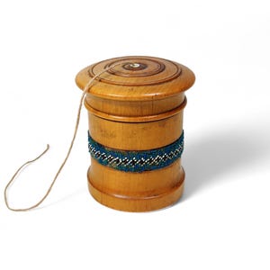 Puede incluir: Una peonza de madera con una cuerda, de forma cilíndrica, adornada con una banda de cuentas azules, blancas y negras. La peonza está hecha de madera clara y tiene un diseño circular.