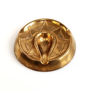 May include: A gold-colored metal ashtray with a decorative design featuring a stylized sunburst and a central indentation.