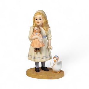 Könnte beinhalten: Porzellanfigur eines jungen Mädchens in einem weißen Kleid, das eine Puppe hält. Das Mädchen steht neben einer weißen Gans mit Hut. Die Figur steht auf einem Holzsockel.