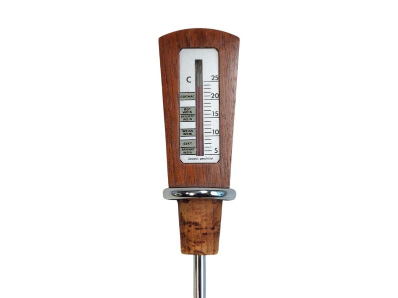 May include: A wine thermometer with a wooden handle featuring a vertical scale from 5 to 25 degrees Celsius. The display includes labels for Cognac, Rotwein, Dessertwein, Weisswein, and Sekt. A silver metal ring separates the handle from a cork stopper.