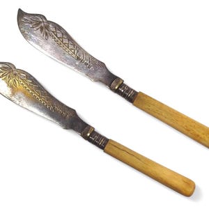 Puede incluir: Dos cuchillos de mantequilla de plata antiguos con diseños dorados ornamentados y mangos de madera.