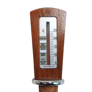 May include: A wooden wine thermometer with a silver-colored metal ring and a cork base. The thermometer features a vertical scale in Celsius with labels for COGNAC, ROT WEIN, DESSERT WEIN, WEISS WEIN, SEKT, and BRANNT WEIN, and the text Gesetzt gesch&uuml;tzt.