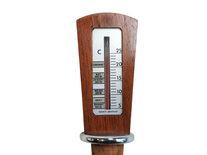 May include: A wooden wine thermometer with a silver-colored metal ring and a cork base. The thermometer features a vertical scale in Celsius with labels for COGNAC, ROT WEIN, DESSERT WEIN, WEISS WEIN, SEKT, and BRANNT WEIN, and the text Gesetzt gesch&uuml;tzt.