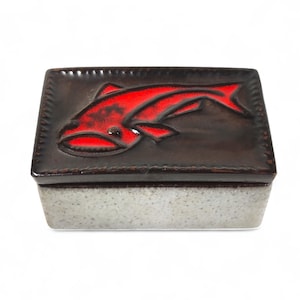 May include: A rectangular trinket box with a dark brown lid featuring a red fish design. The box has a speckled gray base and a dark brown rim. The fish is stylized and centered on the lid.