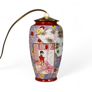 Könnte beinhalten: Dekorative japanische Lampe mit vasenförmigem Körper. Die Lampe hat einen roten Sockel und Rand, einen goldfarbenen Deckel und eine detaillierte bemalte Szene einer Frau in traditioneller Umgebung. Die Lampe hat ein gebogenes, braunes Kabel.