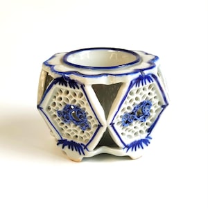 May include: A white ceramic planter with a blue and white floral design. The planter has a hexagonal shape and is open on the sides. It has a small opening at the top for planting.