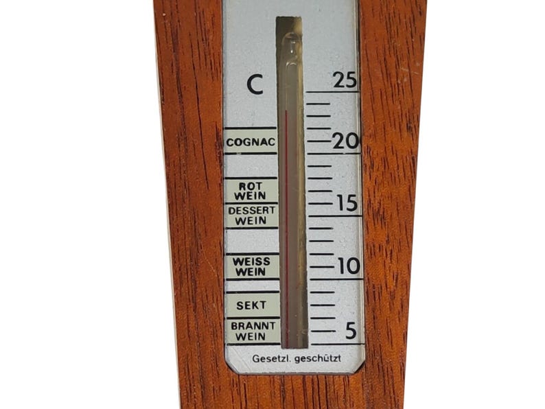 May include: A wooden wine thermometer with a vertical scale in Celsius ranging from 5 to 25 degrees. The white display features black text labels for COGNAC, ROT WEIN, DESSERT WEIN, WEISS WEIN, SEKT, and BRANNT WEIN, with the text Gesetzi. gesch&uuml;tzt at the bottom.