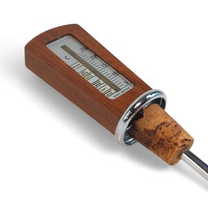 May include: A wine thermometer featuring a rectangular wooden handle with a built-in temperature scale in Celsius, a silver-colored metal ring, a natural cork stopper, and a long, thin stainless steel probe extending from the bottom.