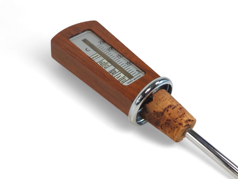May include: A wine thermometer featuring a rectangular wooden handle with a built-in temperature scale in Celsius, a silver-colored metal ring, a natural cork stopper, and a long, thin stainless steel probe extending from the bottom.