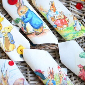 Pode incluir: Guardanapos de tecido branco com ilustrações de Pedro Coelho e outros personagens. Os guardanapos são decorados com botões coloridos em vermelho, amarelo e azul. Dispostos numa cesta de vime.