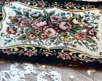 May include: A rectangular tapestry with a black border and a central floral design. The design features a cream-colored background with a detailed arrangement of pink, red, and green flowers, framed by an ornate gold border.