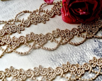 May include: Close-up of delicate, gold-colored lace trim with a floral design. The trim is intricately woven and features a scalloped edge. A deep red rose is visible in the background, adding a touch of color and contrast to the image.