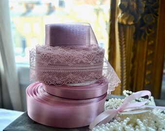 May include: A stack of pink ribbons and lace spools. The bottom spool is a wide, satin ribbon, followed by a lace ribbon, and topped with a sheer ribbon. A string of pearls and a pink ribbon bow are in the foreground.