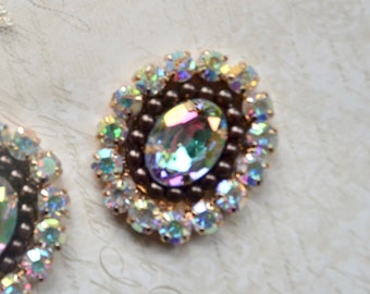 May include: Close-up of two oval-shaped, iridescent rhinestone embellishments. Each rhinestone is surrounded by smaller, clear, iridescent rhinestones and a ring of brown beads. The center stone has a rainbow sheen.