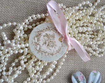 May include: Ivory pearl necklace with a light pink ribbon and a round tag featuring a floral design. The tag is tied with a pink ribbon. The necklace is draped over a textured, neutral-colored surface.