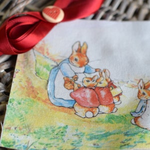 Peut inclure: Un carré de tissu illustré avec une scène de Peter Rabbit. L'image montre une mère lapin et ses enfants, avec Pierre Lapin au premier plan. Un ruban rouge et un bouton en forme de cœur sont en arrière-plan.
