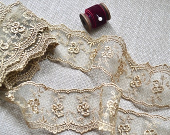 May include: Gold-colored lace trim with floral embroidery and scalloped edges. A spool of burgundy velvet ribbon with a pin is in the background. The lace is partially rolled and spread out, showing the delicate details of the design.