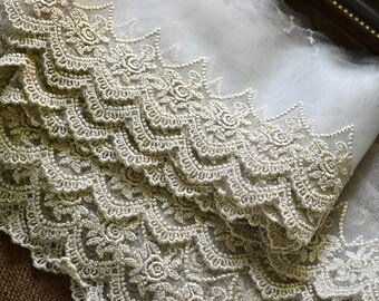 May include: Close-up of a delicate, gold-colored lace trim with a scalloped edge. The lace features intricate floral and scrollwork patterns, creating a luxurious and ornate design. The fabric appears to be a soft, sheer material.