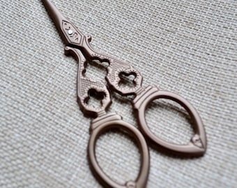 May include: Ornate bronze-colored embroidery scissors with a pointed tip and decorative handles. The scissors have a detailed design with openwork patterns and oval finger holes, ideal for crafting and sewing projects.