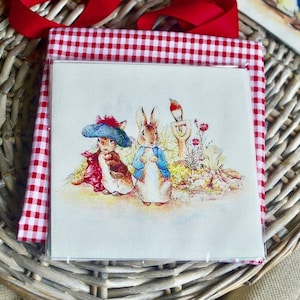 Può includere: Una stampa quadrata con illustrazioni di conigli e un uccello, sullo sfondo di fiori e fogliame. La stampa è racchiusa in una custodia trasparente e incorniciata da un tessuto a quadretti rosso e bianco. È visibile un nastro rosso.