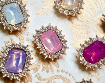 May include: Close-up of several decorative rhinestone embellishments. Each has a rectangular, opalescent center stone in shades of pink, purple, and blue, surrounded by a halo of small clear rhinestones. The settings are gold-toned.