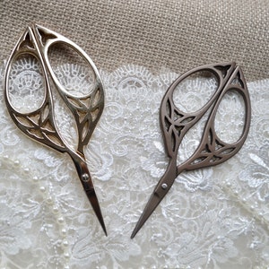 May include: Two decorative scissors with ornate leaf-shaped handles. One is gold-toned, the other is bronze. The scissors are set against a backdrop of lace and burlap, suggesting a craft or sewing theme.